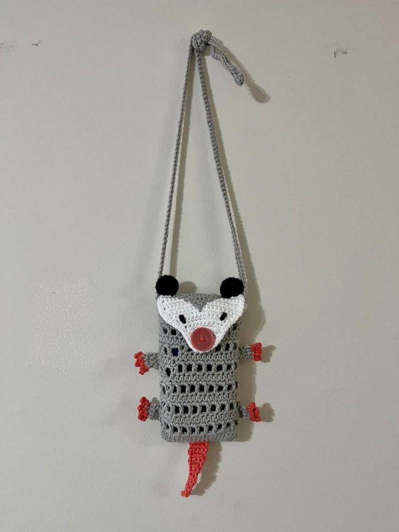 May include: A handmade crochet possum bag in gray with a white face, black ears, and a pink nose and tail. The bag has a gray strap and red feet. The bag is hanging on a white wall.
