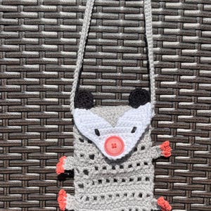 May include: A handmade crochet crossbody bag shaped like an opossum. The bag is gray with a white face, black ears, and coral-colored feet and tail. It features a button closure and a gray strap.