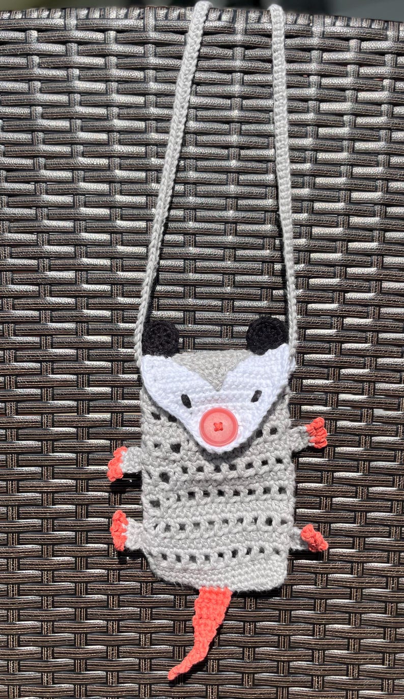 May include: A handmade crochet crossbody bag shaped like an opossum. The bag is gray with a white face, black ears, and coral-colored feet and tail. It features a button closure and a gray strap.