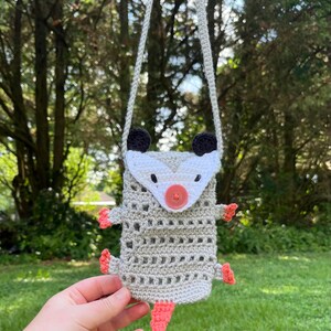 May include: A handmade crochet possum-shaped crossbody bag. The bag is primarily light gray with a white face, black ears, and a coral-colored nose, feet, and tail. It has a button closure and a long, gray strap for wearing over the shoulder.