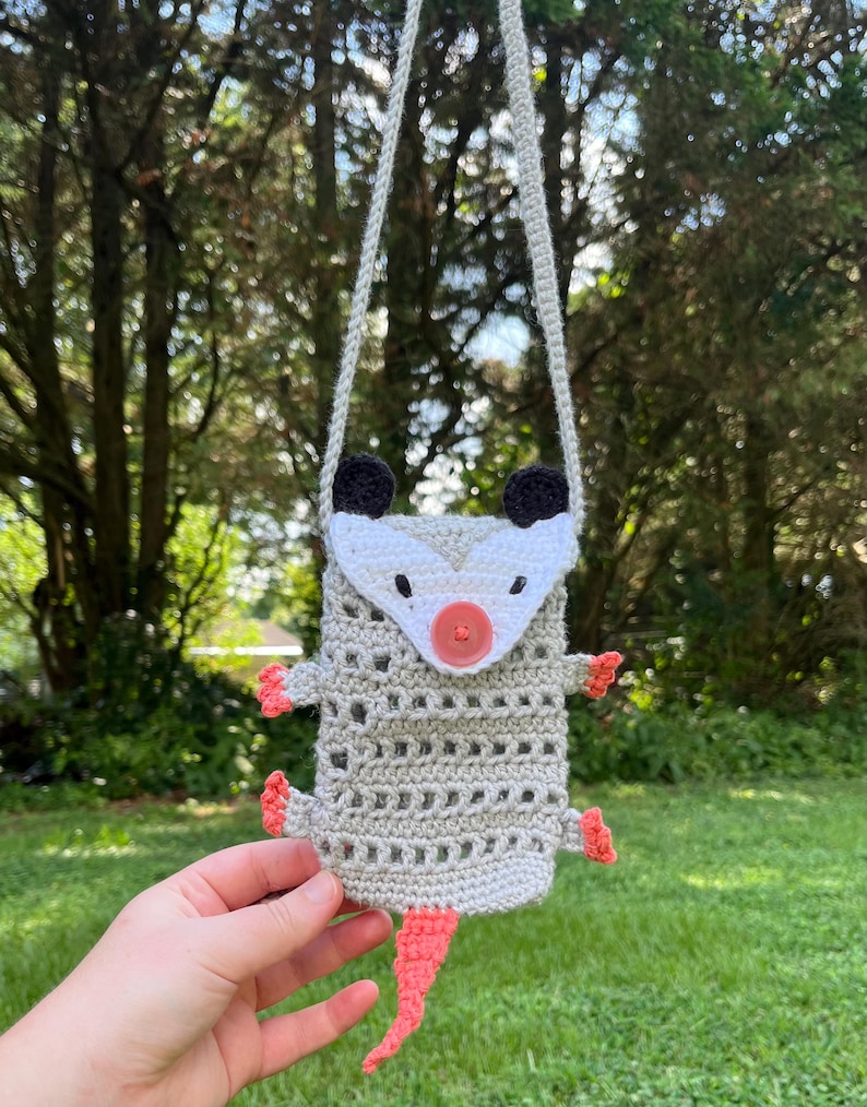 May include: A handmade crochet possum-shaped crossbody bag. The bag is primarily light gray with a white face, black ears, and a coral-colored nose, feet, and tail. It has a button closure and a long, gray strap for wearing over the shoulder.