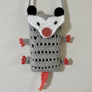 May include: A handmade crochet crossbody bag in the shape of an opossum. The bag is gray with a white face, black ears, and a coral-colored tail and feet. It has a gray strap and a red button closure.
