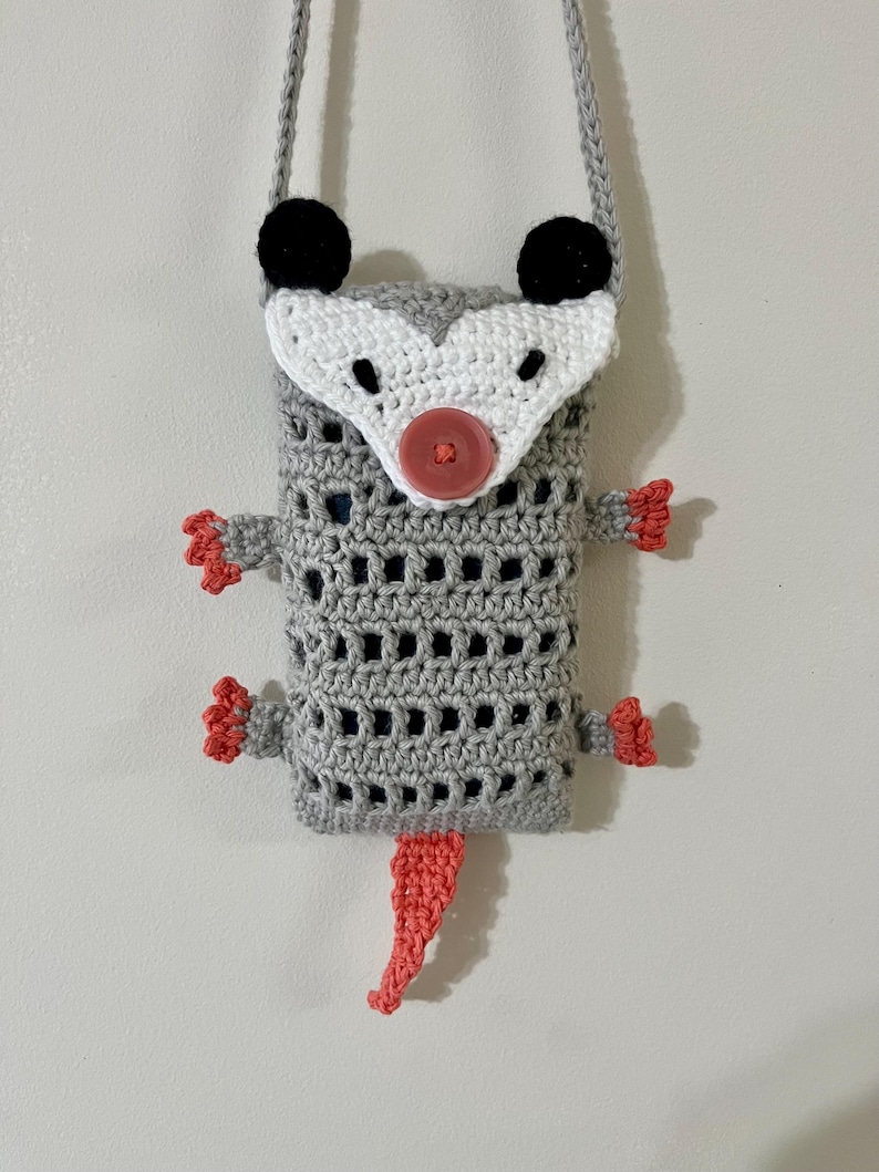 May include: A handmade crochet crossbody bag in the shape of an opossum. The bag is gray with a white face, black ears, and a coral-colored tail and feet. It has a gray strap and a red button closure.