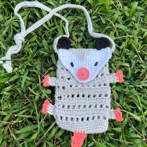 May include: A handmade crochet pouch shaped like an opossum, featuring a gray body, white face, black ears, and a pink nose and tail. The pouch has a gray strap and is set against a backdrop of green grass.