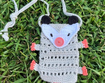 Puede incluir: Bolsa de crochet hecha a mano con forma de zarigüeya, con cuerpo gris, cara blanca, orejas negras y nariz y cola rosas. La bolsa tiene una correa gris y está colocada sobre un fondo de hierba verde.