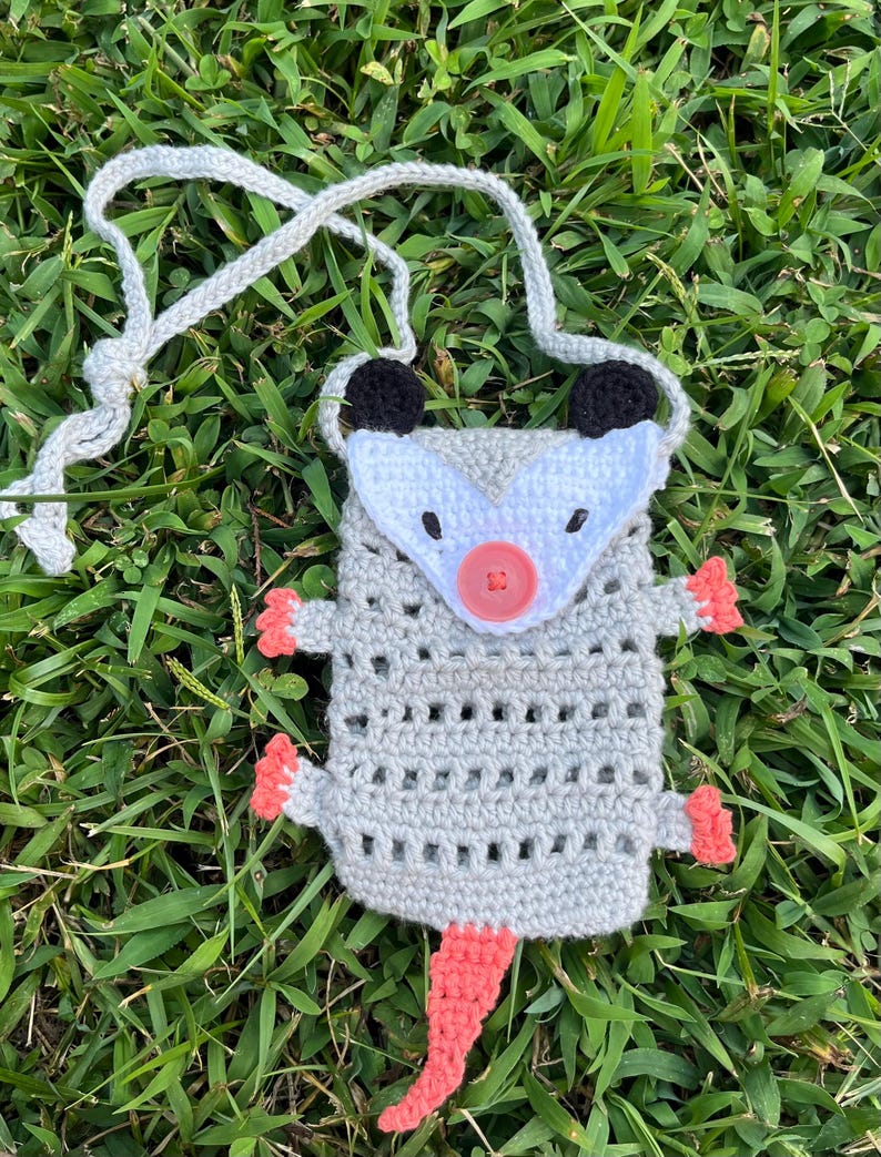 May include: A handmade crochet pouch shaped like an opossum, featuring a gray body, white face, black ears, and a pink nose and tail. The pouch has a gray strap and is set against a backdrop of green grass.
