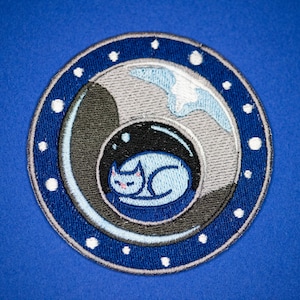 May include: Embroidered patch featuring a sleeping cat inside a bubble, surrounded by a blue and white design. The patch has a circular shape with a blue border and white dots. The cat is light blue with a pink nose.
