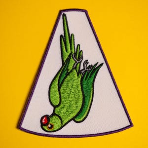 May include: Green embroidered patch of a parrot hanging upside down with its feet in the air. The patch is on a white background with a purple border.
