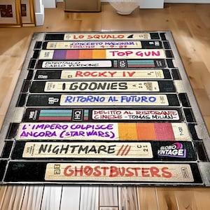 May include: A vintage VHS tape rug featuring titles of popular movies from the 1980s and 1990s, including "Lo Squalo", "Concerto Madonna", "Top Gun", "Rocky IV", "I Goonies", "Ritorno al Futuro", "Delitto al Ristorante Cinese", "L'Impero Colpisce Ancora", "Nightmare", and "Ghostbusters".