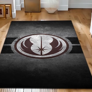 Op de afbeelding: Een zwart en grijs vloerkleed met een verweerde look heeft een wit en grijs Jedi Order-symbool met een bruin lichtzwaard en vleugels. Het symbool wordt omringd door een witte cirkel en twee grijze strepen.