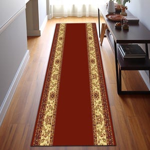Puede incluir: Una alfombra larga y rectangular con un centro rojo oscuro y bordes florales color crema ornamentados. La alfombra está colocada sobre un suelo de madera en un pasillo, añadiendo un toque de elegancia al espacio.