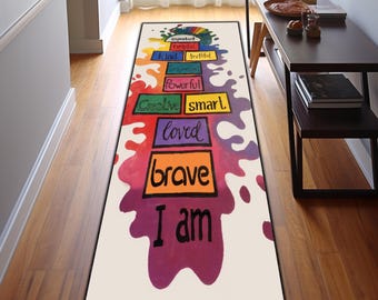 May include: A rectangular white rug with colorful text and paint splatters. The text includes words like "brave," "loved," and "I am." The rug is placed in a hallway with wooden flooring and a white wall.