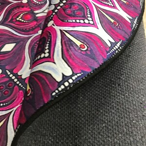 Op de afbeelding: Een ronde yogamat met een mandala-ontwerp in tinten roze, paars en wit. De mat heeft een zwarte rand en een zwarte antislip rug.