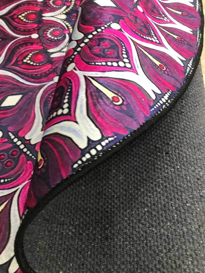 Op de afbeelding: Een ronde yogamat met een mandala-ontwerp in tinten roze, paars en wit. De mat heeft een zwarte rand en een zwarte antislip rug.