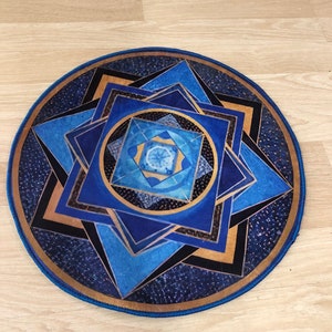 Op de afbeelding: Een rond blauw en gouden mandala-kleed met een geometrisch patroon. Het kleed heeft een blauwe rand en een gouden midden.