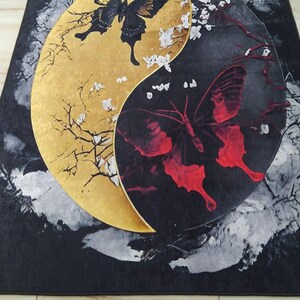 May include: A black and gold rug with a yin yang design featuring a black butterfly on the gold side and a red butterfly on the black side. The butterflies are surrounded by white flowers and branches.