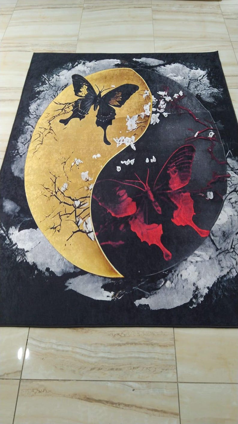 May include: A black and gold rug with a yin yang design featuring a black butterfly on the gold side and a red butterfly on the black side. The butterflies are surrounded by white flowers and branches.