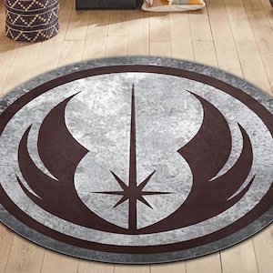 May include: A round, gray and brown rug with a distressed, metallic look. The rug features a stylized symbol of a star with wings, reminiscent of the Rebel Alliance symbol from Star Wars.