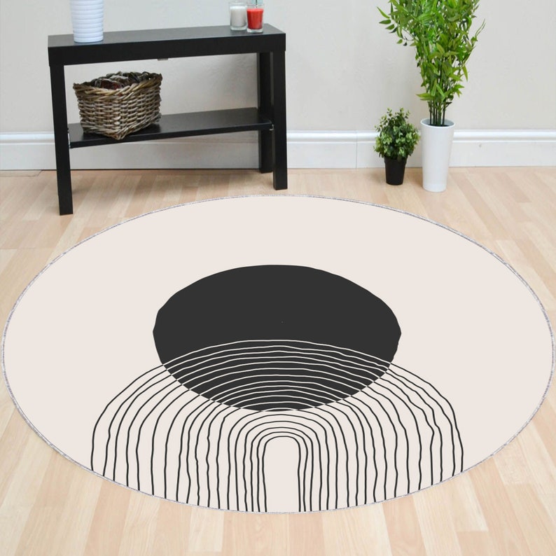 May include: A round cream-colored rug with a black and white abstract design. The design features a large black circle with a series of thin white lines forming an arch shape.