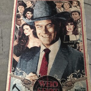 May include: A vintage poster with a central image of a man in a hat and suit, surrounded by other people. The poster displays the text "WHO SHOT J.R.?" and "DALLAS". The design has a faded, distressed appearance with a muted colour scheme.