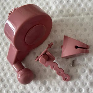 May include: A pink plastic tool with a round base, a handle, and a small, separate piece. The tool has a gear-like mechanism on the round base. The tool is disassembled and laid out on a white surface.
