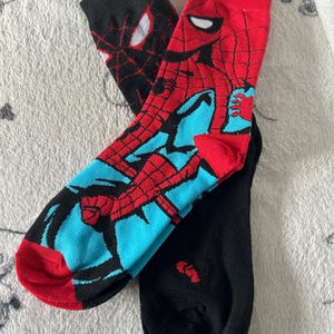May include: Pair of novelty socks featuring Spider-Man. One sock is primarily black with a Spider-Man face design, while the other is red, blue, and black with a full Spider-Man graphic. The socks have red toes and cuffs.