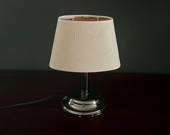 May include: A small table lamp with a cream lampshade, patterned with a repeating design. The lamp has a chrome base and stem. It sits on a dark brown surface. A black cord extends from the lamp.