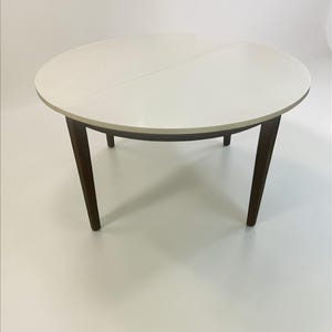 Könnte beinhalten: Ein runder Esstisch mit weißer Tischplatte und dunkelbraunen Beinen. Der Tisch hat eine geteilte Tischplatte und die Beine sind abgewinkelt. Der Tisch hat einen Durchmesser von ca. 102 cm.