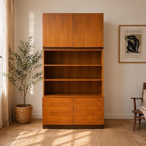 Puede incluir: Un mueble alto de madera con un acabado c&aacute;lido de color marr&oacute;n rojizo. El mueble tiene dos puertas superiores, estanter&iacute;as abiertas en el medio y dos puertas inferiores. Un grabado enmarcado de Matisse cuelga en la pared. Un peque&ntilde;o &aacute;rbol en una cesta tejida est&aacute; cerca. El mueble mide unos 180 cm de alto.