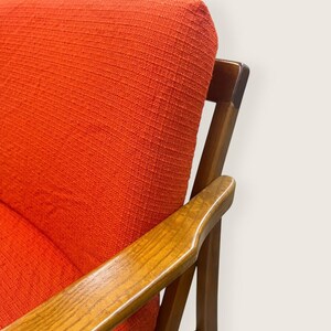 May include: Close-up of a mid-century modern armchair. The chair features a vibrant orange fabric upholstery and wooden armrests. The wooden frame has a warm, natural finish. The design is simple and elegant, showcasing a retro aesthetic.