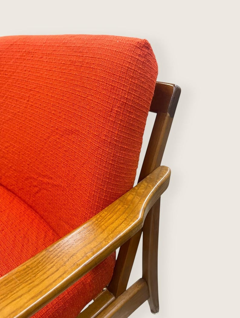 May include: Close-up of a mid-century modern armchair. The chair features a vibrant orange fabric upholstery and wooden armrests. The wooden frame has a warm, natural finish. The design is simple and elegant, showcasing a retro aesthetic.
