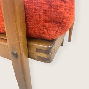 May include: Close-up of a mid-century modern chair. The chair features a wooden frame with a reddish-orange upholstered cushion. The wood has a warm, natural finish. The joinery is visible, showcasing the craftsmanship. The chair's design is simple and elegant.