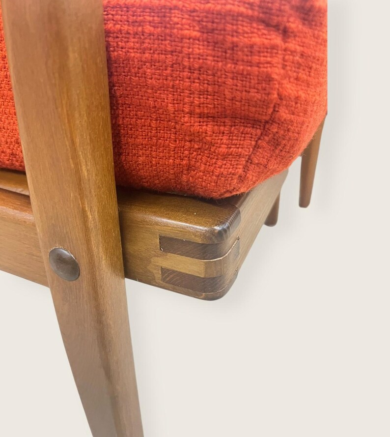 May include: Close-up of a mid-century modern chair. The chair features a wooden frame with a reddish-orange upholstered cushion. The wood has a warm, natural finish. The joinery is visible, showcasing the craftsmanship. The chair's design is simple and elegant.