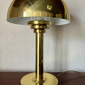 Puede incluir: L&aacute;mpara de escritorio dorada con una pantalla en forma de c&uacute;pula y una base cil&iacute;ndrica. La l&aacute;mpara tiene un acabado pulido y un interior blanco en la pantalla. La l&aacute;mpara est&aacute; sobre una superficie marr&oacute;n oscuro. Altura aproximada 30 cm.