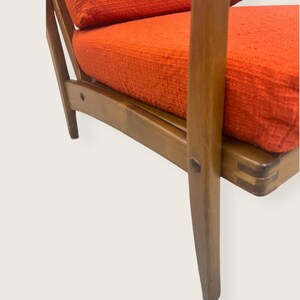 May include: Close-up of a mid-century modern armchair. The chair features a wooden frame with a warm, brown finish. The seat and back cushions are upholstered in a textured, bright orange fabric. The chair's design showcases clean lines and angled legs.