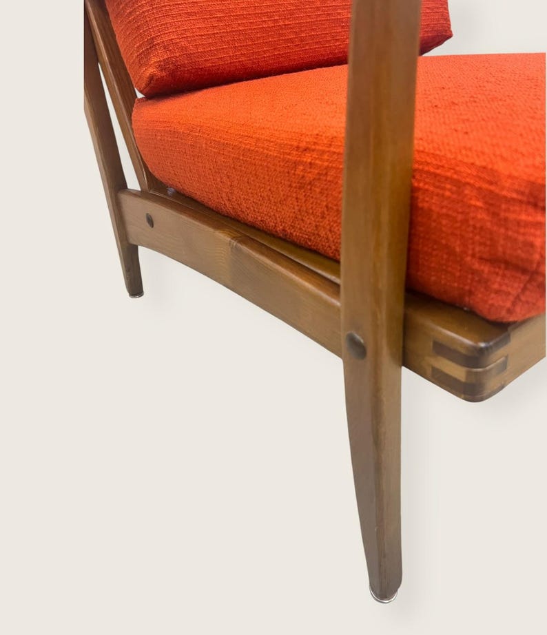 May include: Close-up of a mid-century modern armchair. The chair features a wooden frame with a warm, brown finish. The seat and back cushions are upholstered in a textured, bright orange fabric. The chair's design showcases clean lines and angled legs.