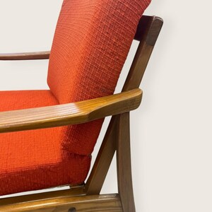 May include: Mid-century modern armchair with a vibrant orange fabric seat and backrest. The chair features a wooden frame with armrests, showcasing a classic design. The wood has a warm, natural finish, complementing the bold color of the upholstery.