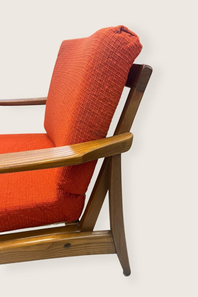 May include: Mid-century modern armchair with a vibrant orange fabric seat and backrest. The chair features a wooden frame with armrests, showcasing a classic design. The wood has a warm, natural finish, complementing the bold color of the upholstery.