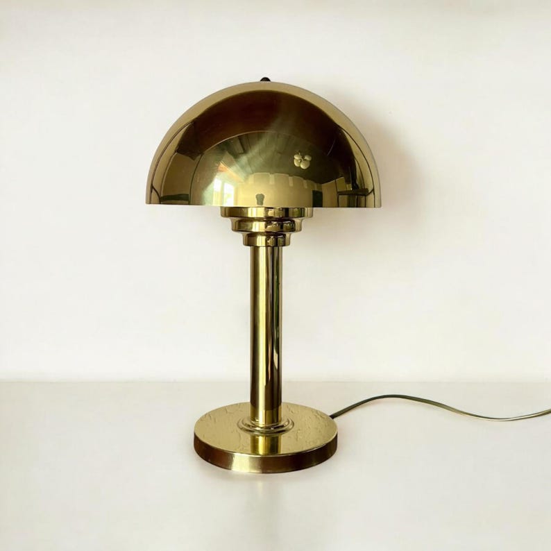 Puede incluir: Una l&aacute;mpara de mesa de lat&oacute;n con una pantalla en forma de c&uacute;pula y una base cil&iacute;ndrica. La l&aacute;mpara tiene un acabado pulido, que refleja el entorno. El dise&ntilde;o recuerda la est&eacute;tica moderna de mediados de siglo. La l&aacute;mpara est&aacute; conectada a una fuente de alimentaci&oacute;n por un cable negro.