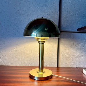 Puede incluir: Una l&aacute;mpara de escritorio de lat&oacute;n con una pantalla en forma de c&uacute;pula de color oscuro. La l&aacute;mpara tiene una base y un v&aacute;stago cil&iacute;ndricos, y una base circular. La l&aacute;mpara est&aacute; sobre una superficie de madera.