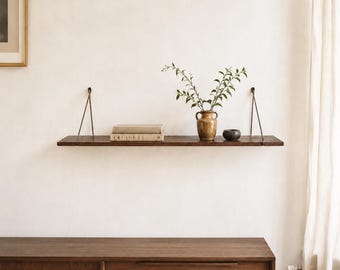 Könnte beinhalten: Ein dunkelbraunes Wandregal aus Holz, das an zwei dünnen Metallstangen aufgehängt ist. Auf dem Regal liegen zwei beige Bücher, eine hellbraune Keramikvase mit grünen Blattzweigen und eine kleine dunkelgraue Schale. Es hängt über einem Holzschrank.