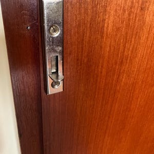 Puede incluir: Primer plano de una puerta de madera con una cerradura corredera plateada brillante. La puerta es de color marr&oacute;n rojizo c&aacute;lido, y la cerradura tiene un ojo de cerradura y un mecanismo deslizante.