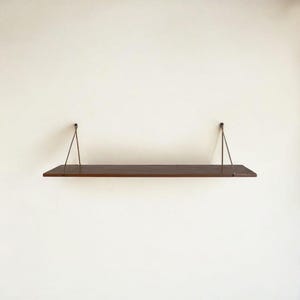 May include: A dark brown wooden floating shelf suspended by metal wire supports. The rectangular shelf is mounted on a white wall, showcasing a minimalist design. The shelf is approximately 36 inches long.