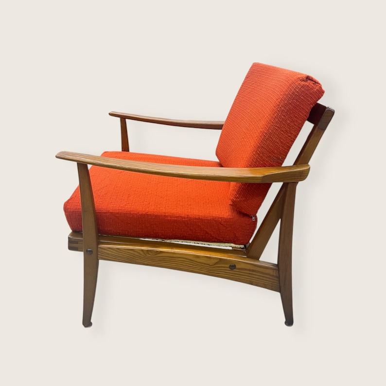 May include: Mid-century modern armchair with a wooden frame and vibrant orange cushions. The chair features a low profile, angled legs, and curved armrests. The cushions are a textured fabric.