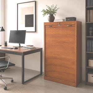 Könnte beinhalten: Ein hoher, rechteckiger Holzschrank mit einer vertikalen Rollladentür und zwei Metallgriffen an der Oberseite. Der Schrank hat eine warmbraune Holzmaserung und einen zentralen Schlossmechanismus mit Schlüsselloch und steht aufrecht im Raum.
