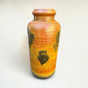 May include: A tall, cylindrical ceramic vase with a textured surface. The vase features a gradient of colors, transitioning from yellow to orange and brown. Dark, abstract shapes are scattered across the surface. The vase has a narrow neck and a wide base.