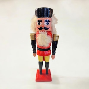 Op de afbeelding: Een houten notenkrakerfiguur met een zwarte hoed, een rode jas en een gele broek. Het figuur heeft een witte baard en snor, met gouden accenten op de armen. Het staat op een rode basis.