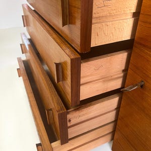 Puede incluir: Una c&oacute;moda de madera con cuatro cajones, algunos parcialmente abiertos. La c&oacute;moda est&aacute; hecha de madera marr&oacute;n clara y oscura, con vetas visibles. Los cajones tienen tiradores rectangulares y marcas manuscritas. El lateral tiene una cerradura de metal.