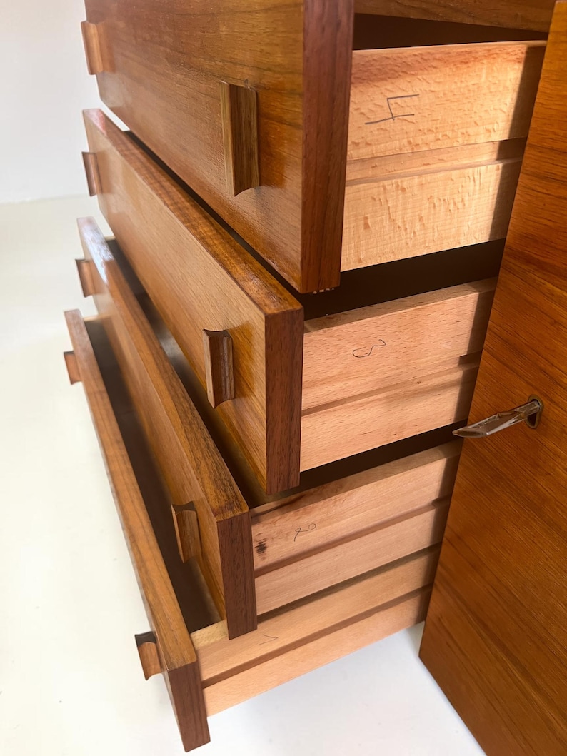 Puede incluir: Una c&oacute;moda de madera con cuatro cajones, algunos parcialmente abiertos. La c&oacute;moda est&aacute; hecha de madera marr&oacute;n clara y oscura, con vetas visibles. Los cajones tienen tiradores rectangulares y marcas manuscritas. El lateral tiene una cerradura de metal.
