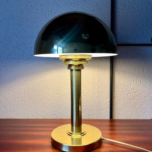 Puede incluir: Una l&aacute;mpara de escritorio de estilo vintage con una pantalla negra en forma de c&uacute;pula y una base y un v&aacute;stago de lat&oacute;n. La l&aacute;mpara est&aacute; encendida, emitiendo un brillo c&aacute;lido. La l&aacute;mpara est&aacute; sobre una superficie de madera oscura.